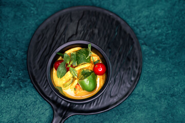 Thai coconut based green curry with vegetables like green bell pepper, red bell pepper, baby corn, cherry tomato, kaffir lime leaves, served with rice in a black platter against a green background