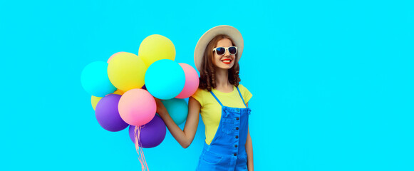 Obraz na płótnie Canvas Summer image of happy laughing young woman with bunch of colorful balloons having fun wearing straw hat on blue studio background