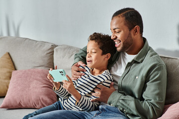 happy african american father and son in casual outfits sitting on sofa looking at mobile phone