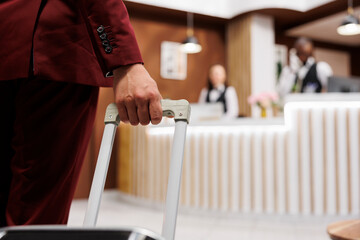 White collar worker with luggage arriving at hotel reception lobby, preparing to see room reservation. Young adult travelling on business meetings, carrying suitcase internationally. Close up.