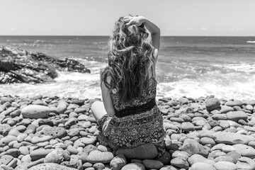 Girl with blond long hairs sitting on a pebble beach in Fuerteventura, Canary Islands, Spain, looking at the sea ocean photo from behind