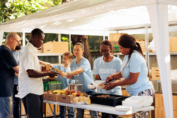 Individuals of diverse backgrounds gather outdoors to give donated food and non-perishables, providing support to the hungry and homeless. Multiracial volunteers share fresh free meals to poor people.