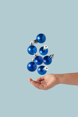 Fototapeta premium Shiny Blue Christmas decoration balls falling down into hand isolated on pastel blue background. Blue Christmas aesthetics.