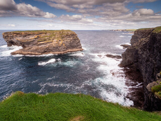 Fototapeta na wymiar View on rough small island in the ocean from a cliff, Ireland, Kilkee area. Warm sunny day, blue cloudy sky. Travel, tourism and sightseeing concept. Irish landscape and coastline.