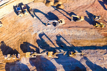 Heavy dump trucks, bulldozers, and excavators on yellow clay construction site. Long shadows on the ground. Top view at sunset.