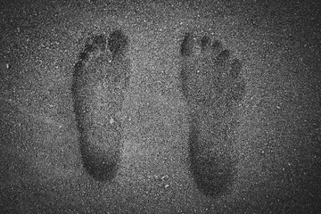 Black and white human barefoot footprints in wet, black volcanic sand of a beach. 