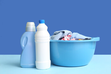 Plastic basin with dirty clothes and bottles of laundry detergent on blue background