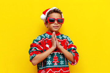 african american boy in a christmas sweater and santa hat and glasses folded his hands in front of...