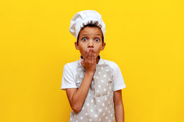 shocked african american boy in chef's uniform and hat covers his mouth with his hand and looks at...