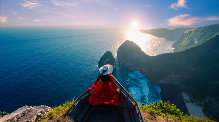 Woman in red dress at Kelingking Beach viewpoint with the light of the sun shining at sunset On the island of Nusa Penida, Indonesia - obrazy, fototapety, plakaty