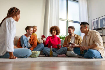 multiracial group of young friends playing cards drinking beer and having fun at home, students sitting on the floor