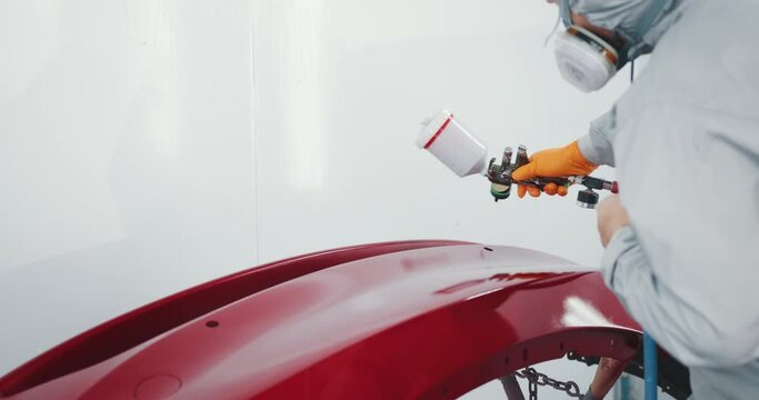 Man Working in Car Service Dressed in Special Uniform and Mask Paints White Auto Bumper Red Using Airless Spray Gun in Booth Closeup. Worker Painting a Red Electric Car in Special Garage