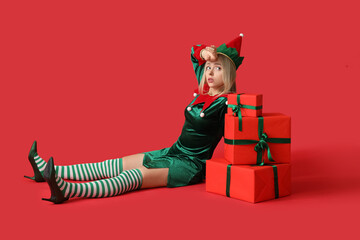Tired woman in elf costume with Christmas gift boxes on red background - obrazy, fototapety, plakaty