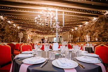 Close-up photo of chromium candlestick with five unlit white wax candles placed on the table prepared for wedding reception. Round tables with red chairs