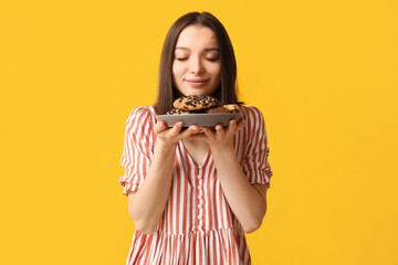 Beautiful young woman with plate of tasty cookies on yellow background