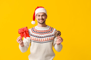 Handsome man in Santa hat with gift box and tasty cookie on yellow background