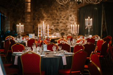 Close-up photo of chromium candlestick with five unlit white wax candles placed on the table prepared for wedding reception. Round tables with red chairs