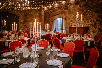 Close-up photo of chromium candlestick with five unlit white wax candles placed on the table prepared for wedding reception. Round tables with red chairs