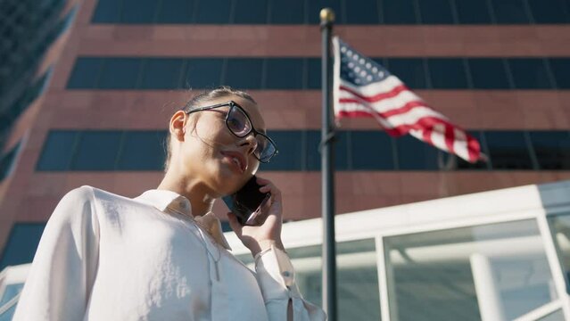 Smiling woman having phone call under large American Flag flattering at wind in golden sunshine at business office in city downtown. Politics, Economy, Business Concept.Low angle cinematic slow motion