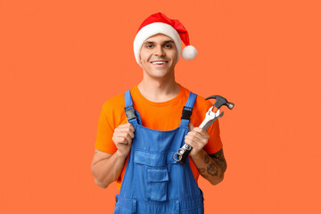 Male plumber in Santa hat with hammer and wrenches on orange background