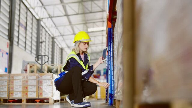 Young caucasian woman warehouse workers holding digital tablet checking inventory management packaging boxes. Woman staff wearing vest and safety helmet and walking count the box at storehouse