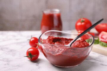 Glass bowl of tasty tomato paste with spoon and ingredients on white marble table, closeup. Space for text