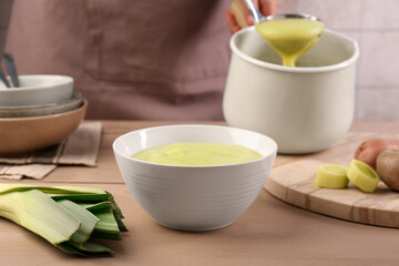 Woman with ladle pouring tasty leek soup at wooden table, focus on bowl