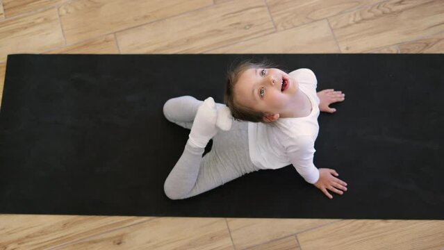 Top view of preschool girl in sports wear is engaged in gymnastics at home.Sporty cute female sitting on mat and stretching by touching tips of legs to head in bright spacious room , copy space.