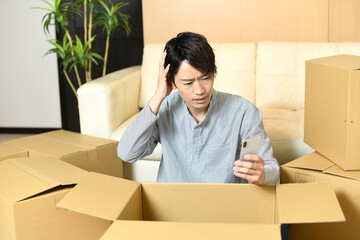 段ボールの前でスマホを持ちながら困るアジア人の男性