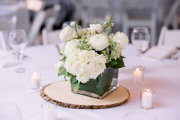 White hydrangeas in a glass square vase sitting on a wood saucer surrounded by candles on a white tablecloth table.