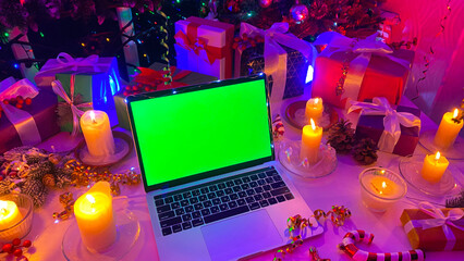 Laptop with a chroma key on the screen is lying on the Christmas table. Packed gift boxes, burning...
