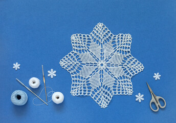 Winter crafts and handmade gifts concept. Hand crocheted lace doily in shape of Christmas snowflake on blue background with skeins of cotton yarn and crochet hooks. Flat lay, copy space, mock up - obrazy, fototapety, plakaty
