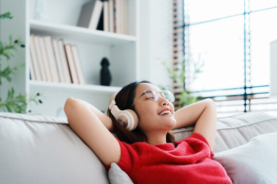 Relaxed Young Asian Woman Enjoying Rest On Comfortable Sofa, Calm Attractive Girl Relaxing On Couch, Breathing Fresh Air With Eyes Closed, Wearing Eyeglasses And Headset, Meditating At Home