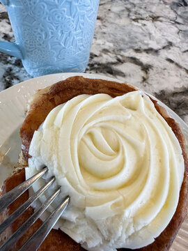 A cinnamon roll with cream cheese frosting sits by a blue embossed coffee mug. A granite countertop is in the background. 