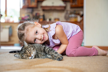 Cute little girl hugging gray cat and lying on carpet in home living room