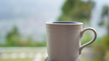 A white cup of coffee or tea object on the table with blur green garden outdoor background. Empty cup for inspirational concept and any creative background. Morning hot tea or coffee backgrounds.