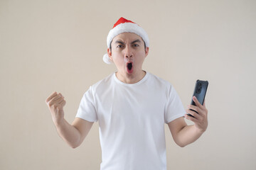 Young Asian Santa man is excited while holding the smartphone and looking at the camera. Isolated beige background. Christmas concept.