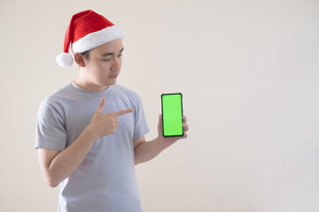 Young Asian Santa man is pointing at his green screen smartphone. Isolated beige background. Christmas concept.