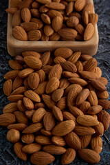 Delicious roasted almonds close-up on a gray textured background are scattered from a square-shaped wooden plate. Raw peeled almonds stacked in a heap on the table, top view