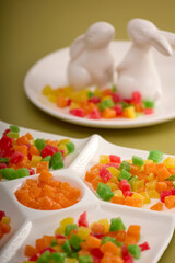 Delicious multi-colored candied fruit is laid out on a white curly dish on a yellow background. In a white porcelain plate with figures of hares, a dessert of sweet candied fruit is laid out