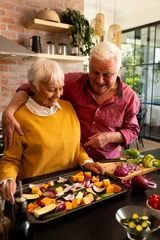 Tuinposter Happy caucasian senior couple preparing fresh vegetables, embracing and talking in kitchen © WavebreakMediaMicro