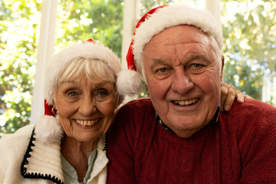 Happy caucasian senior couple in santa hats having christmas video call, smiling in sunny room