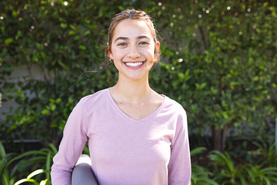 Portrait of happy biracial woman holding yoga mat smiling in sunny garden