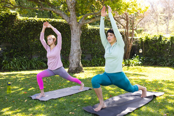 Focused biracial lesbian couple practicing yoga standing in lunge pose in sunny garden
