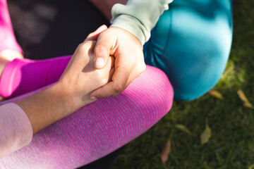 Midsection of biracial lesbian couple practicing yoga sitting holding hands in garden