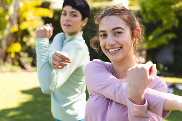 Happy biracial lesbian couple practicing yoga standing and stretching arms in sunny garden