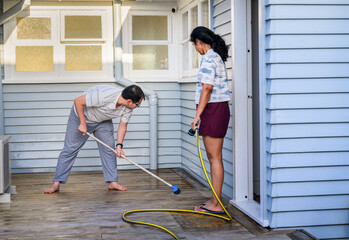 Couple cleaning wooden deck with brush and water sprayer.