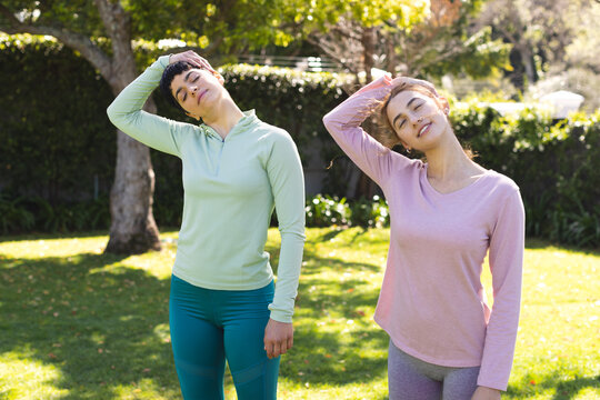 Happy biracial lesbian couple practicing yoga standing and stretching necks in sunny garden