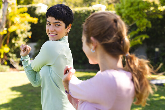 Happy biracial lesbian couple practicing yoga standing and stretching arms in sunny garden