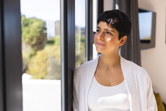 Thoughtful biracial woman looking out of window at home smiling on sunny day, copy space